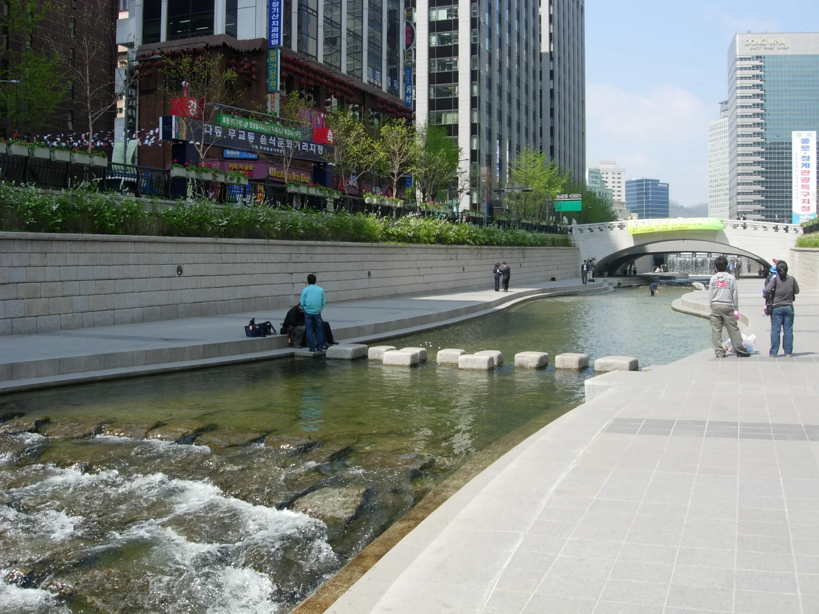 A view from the Cheonggyecheon walking path showing people, stepping stones, flowing water and a stone bridge